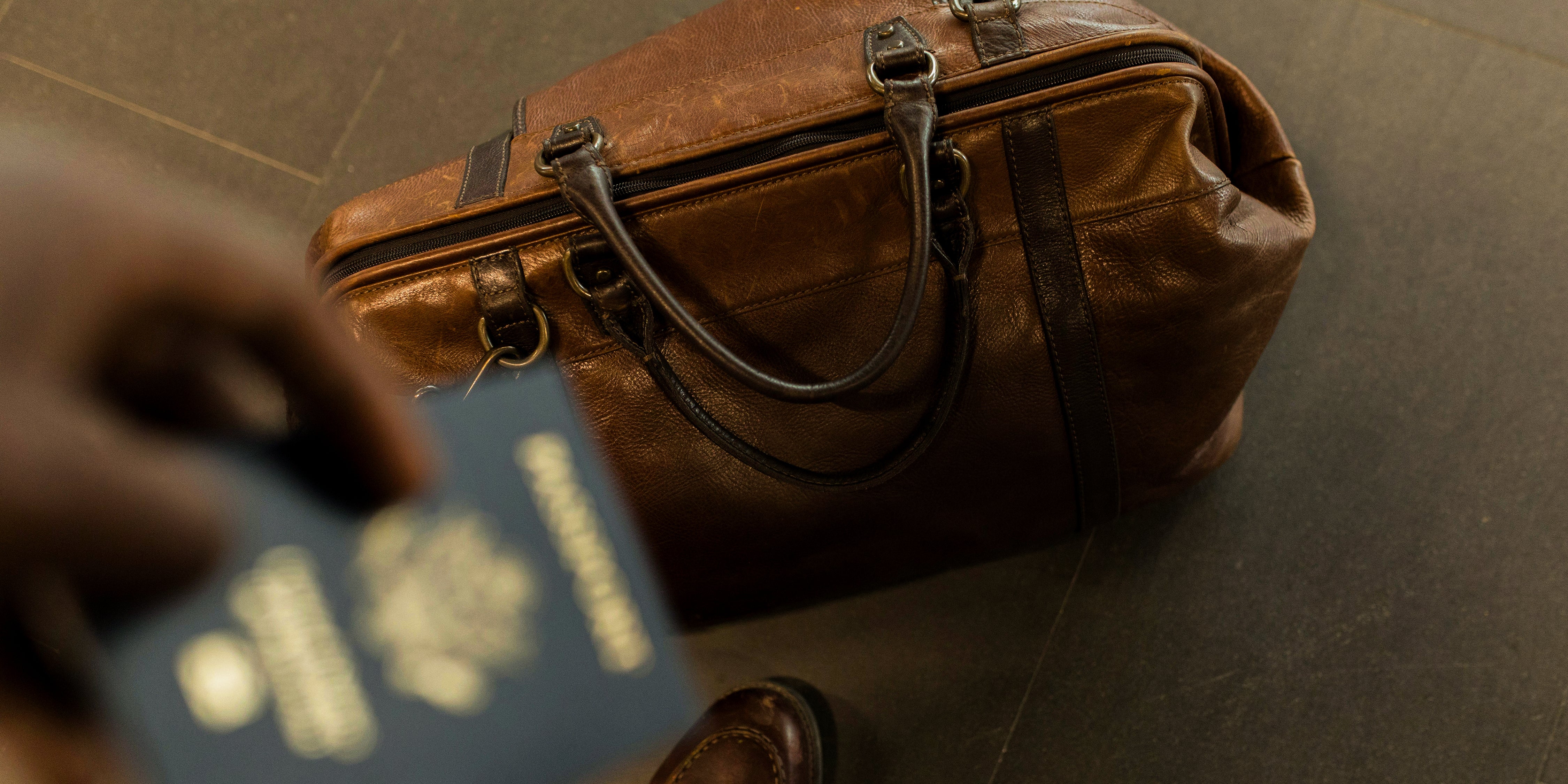 Close-up of a brown leather travel bag on a tile floor, with a hand holding a U.S. passport in the foreground.